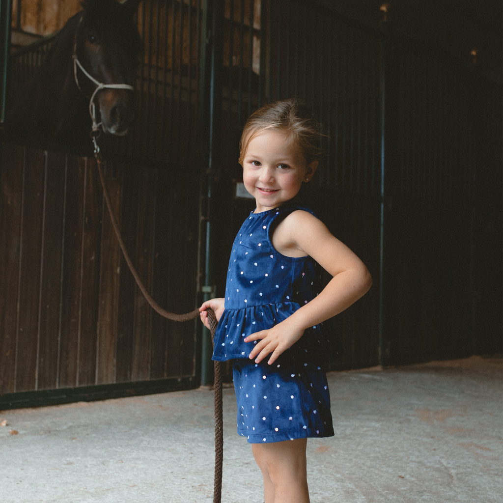 Little girl smiling in a playful Piglo Kids navy outfit with sequins,standing next to a horse in a stable.