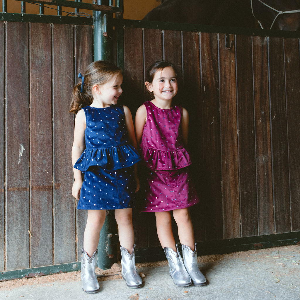 Two little girls smiling in playful Piglo kids outfits with sequins, one in navy blue and the other in red, wearing shiny silver cowboy boots